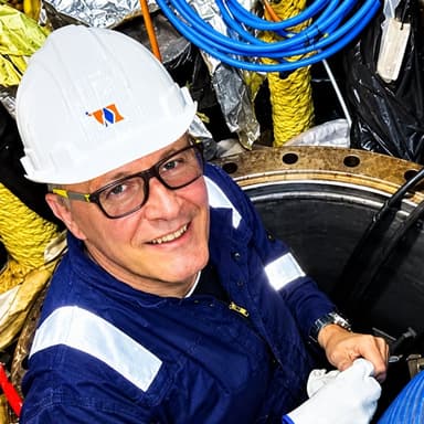 Gerhard Hoffmann in a hard hat in a cable shaft aboard ship, smiling, with work gloves and a hose
